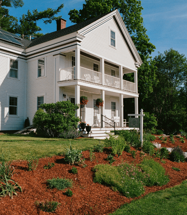 A white two-story house with a front porch and lush garden landscaping.