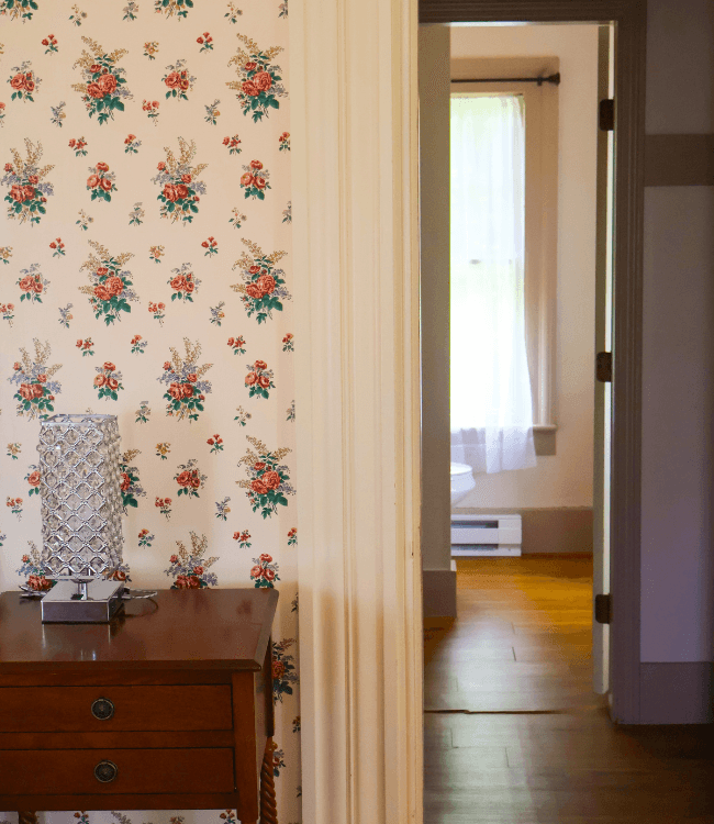 A view into a softly lit room through a doorway, with floral wallpaper and a table lamp in the foreground.