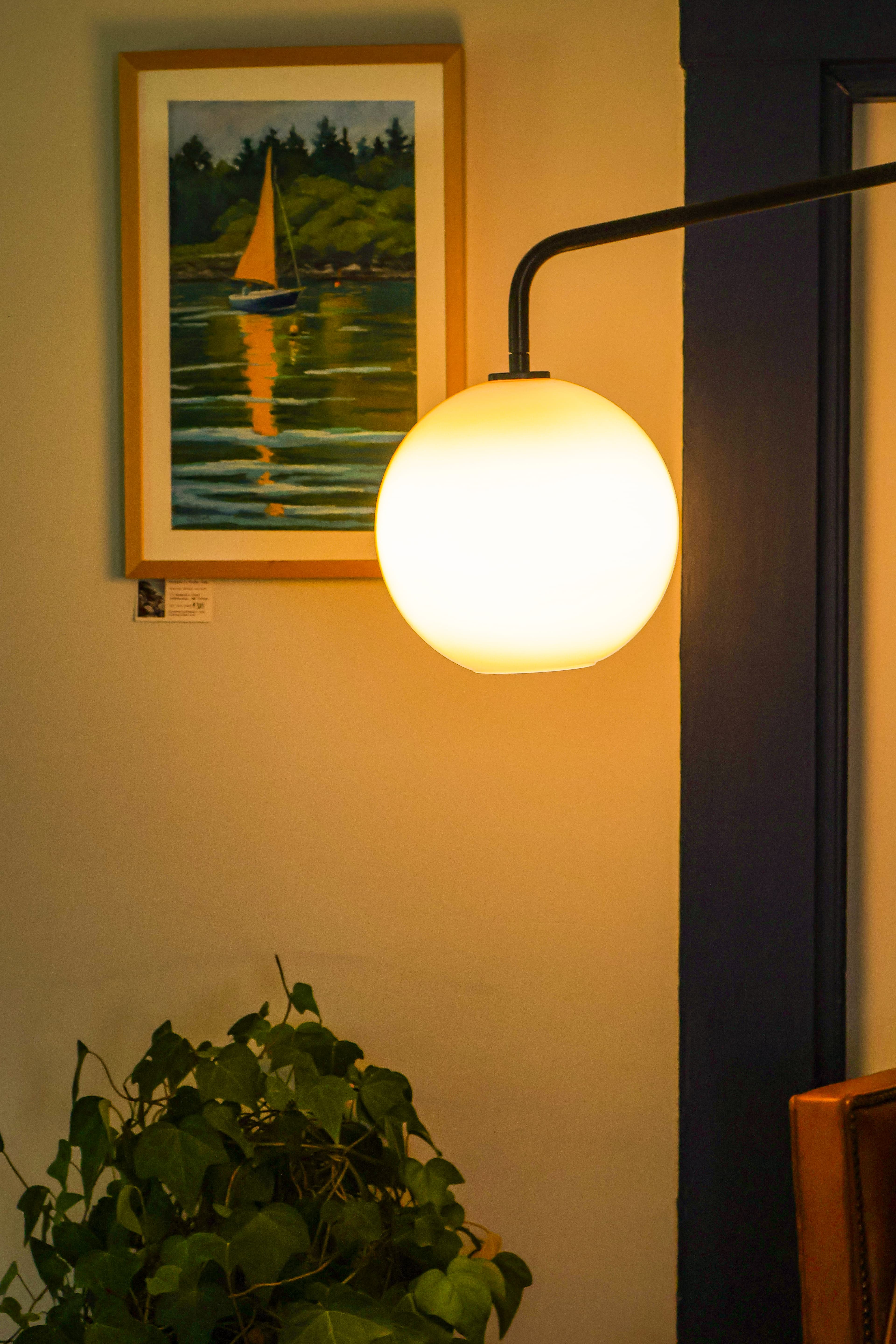 A glowing round lamp hangs beside a framed painting of a sailboat on water, with a green potted plant below.