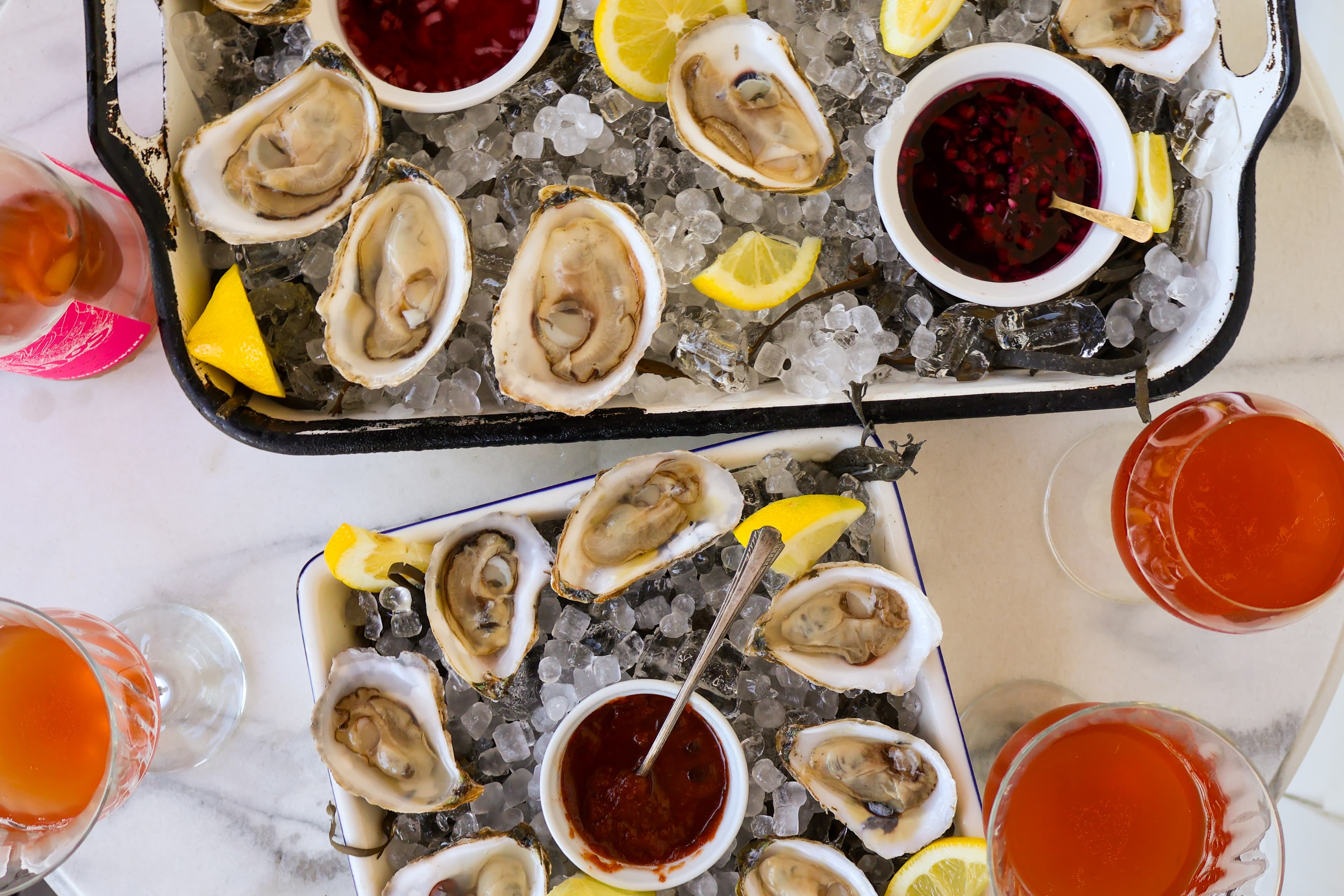 A platter of fresh oysters on ice, garnished with lemon slices and accompanied by sauces and colorful cocktails.