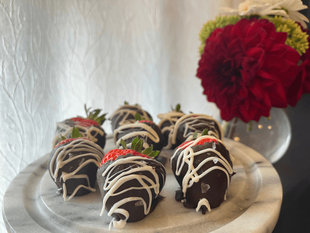 A plate of chocolate-covered strawberries drizzled with white chocolate, next to a vibrant flower arrangement.