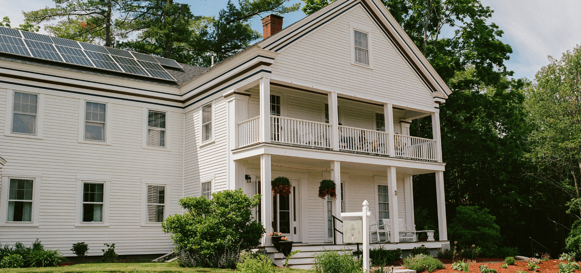 A white two-story house with a porch, solar panels on the roof, and lush greenery around it.