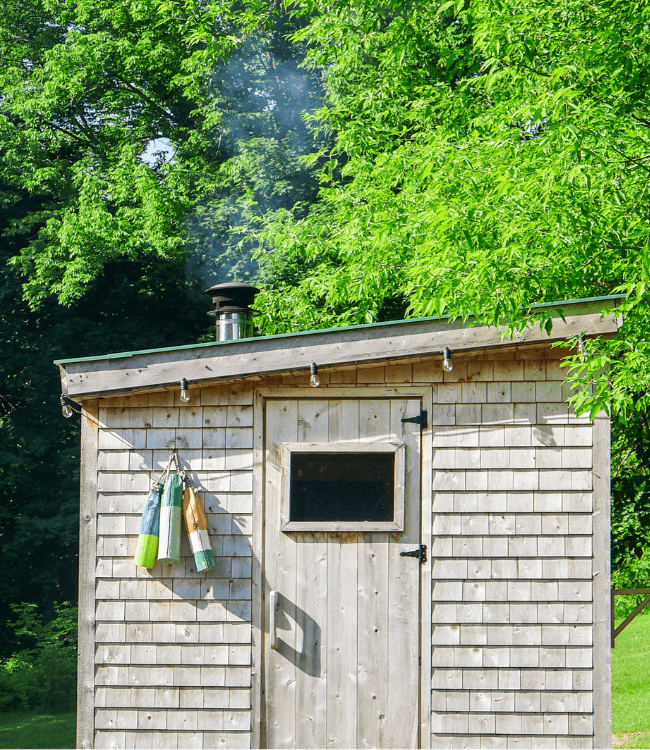 A wooden shed with a chimney and colorful towels hanging on the door, surrounded by green foliage.