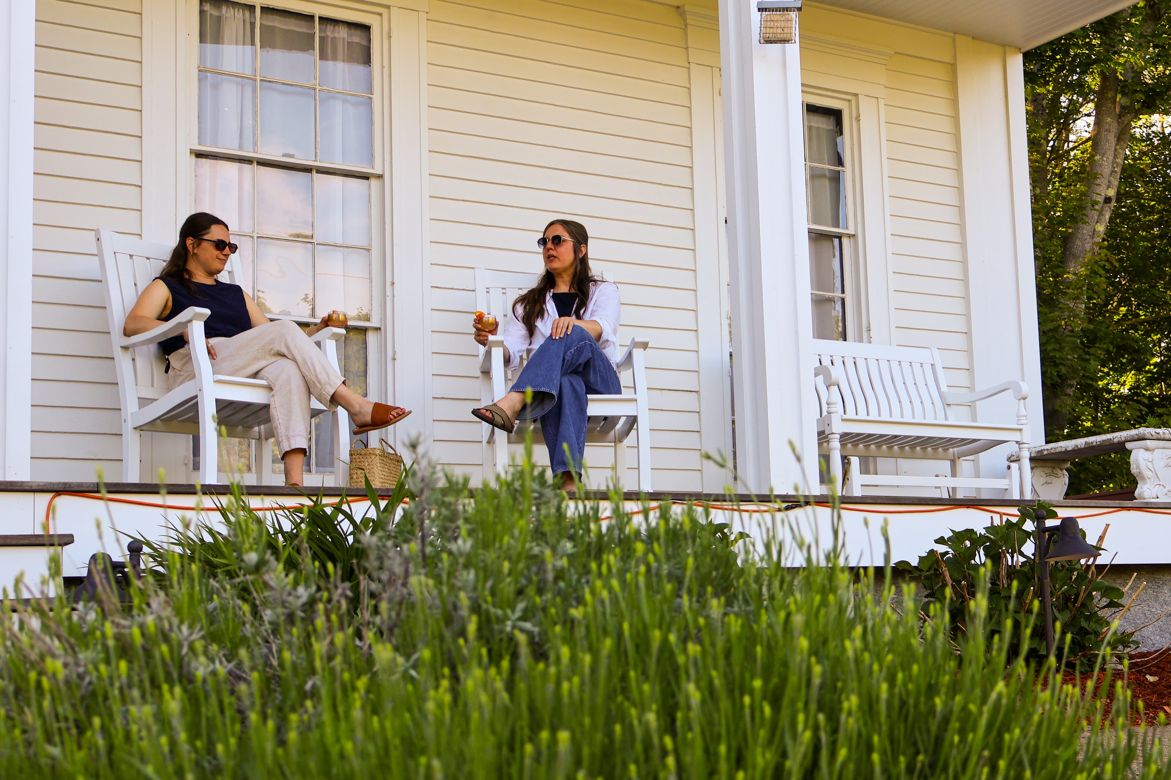 Two women wearing sunglasses relax on a porch, seated on white rocking chairs amid greenery.