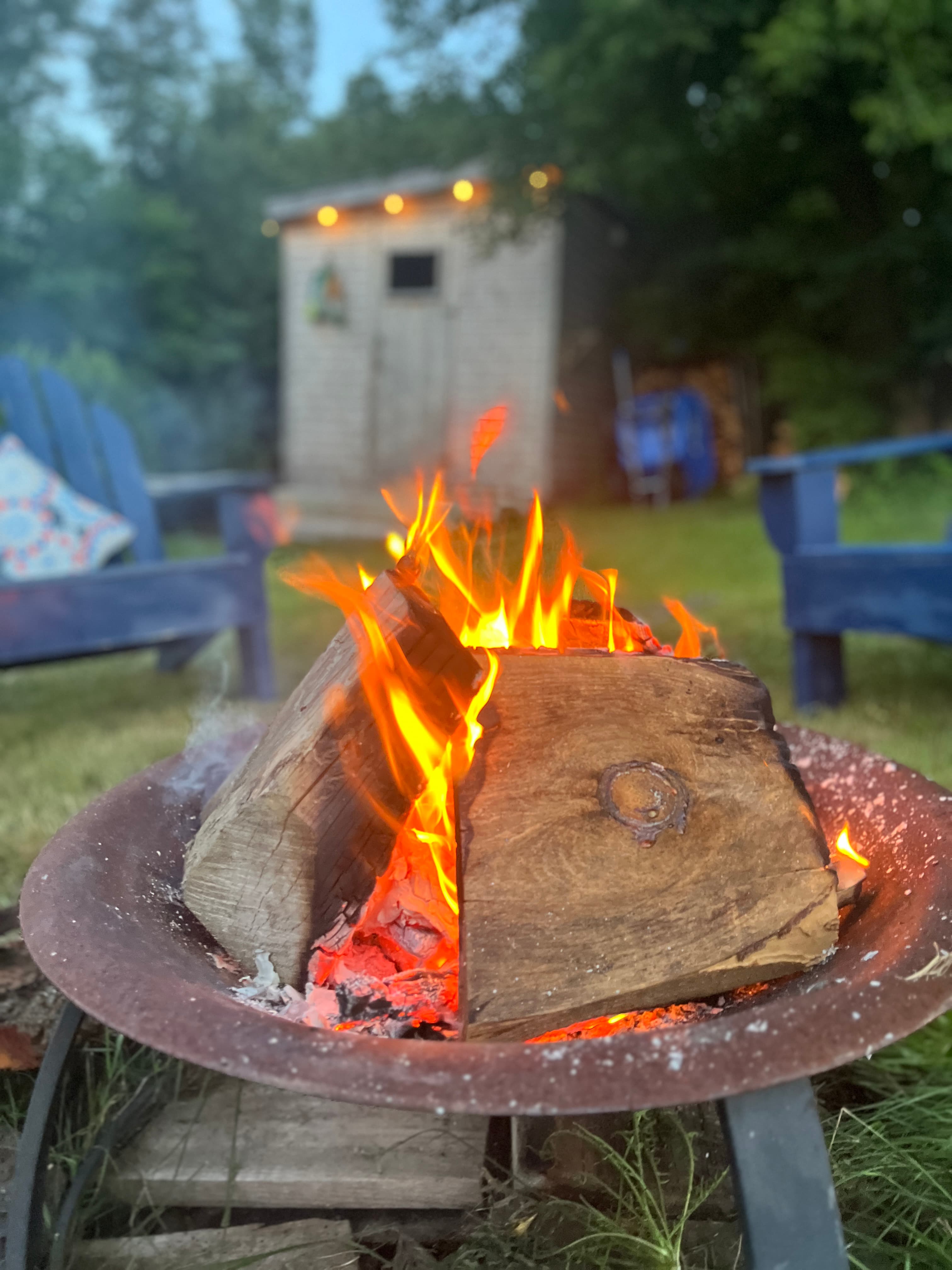 A glowing campfire with logs burning brightly, surrounded by blue seating and a shed in the background.