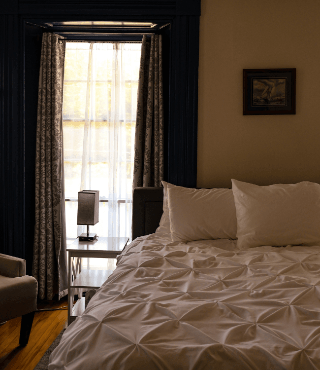Cozy bedroom with a made bed, lamp, and window dressed in sheer curtains.