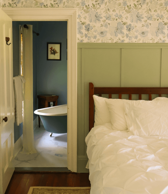 A cozy bedroom featuring a bed with white linens, leading to a bathroom with a vintage bathtub.