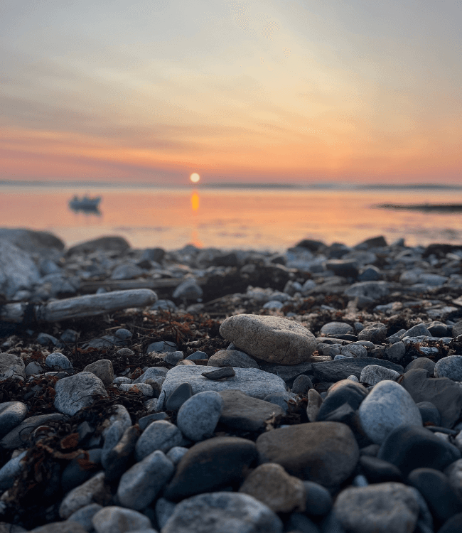 A rocky shore at sunset with a distant boat on calm water.