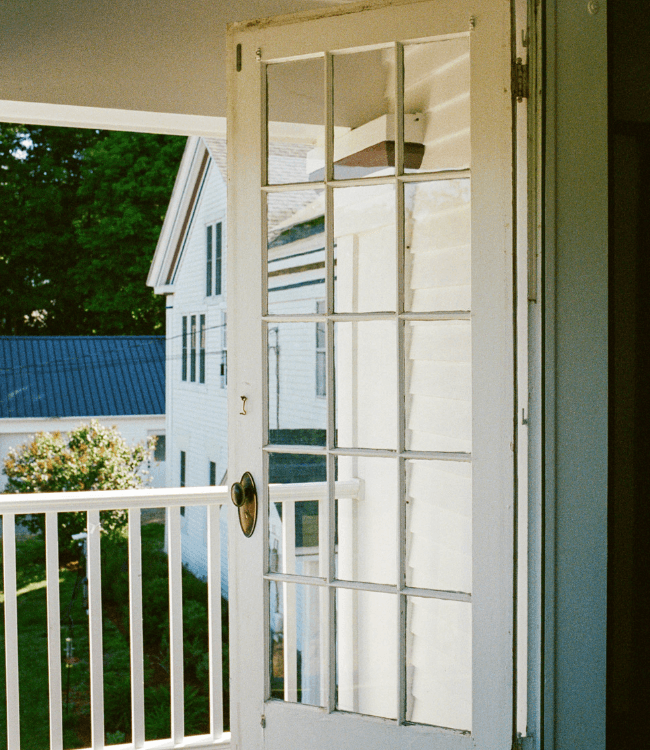 A white, glazed door open to a balcony with views of green trees and neighboring houses.