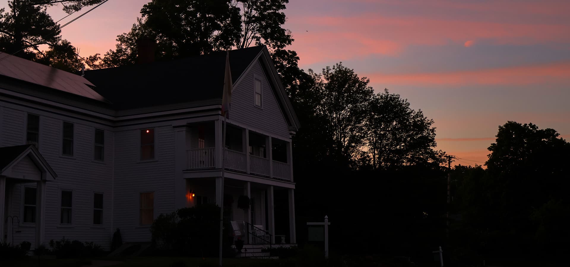 A white house with illuminated windows during sunset, surrounded by trees and a colorful sky.