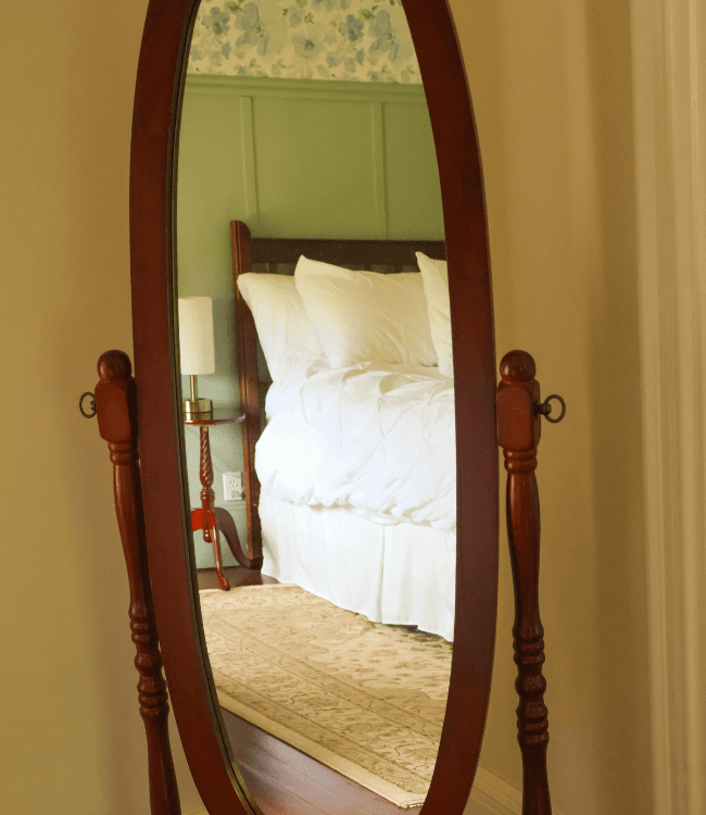 A wooden framed mirror reflecting a bedroom with a bed and lamp against a green wall.