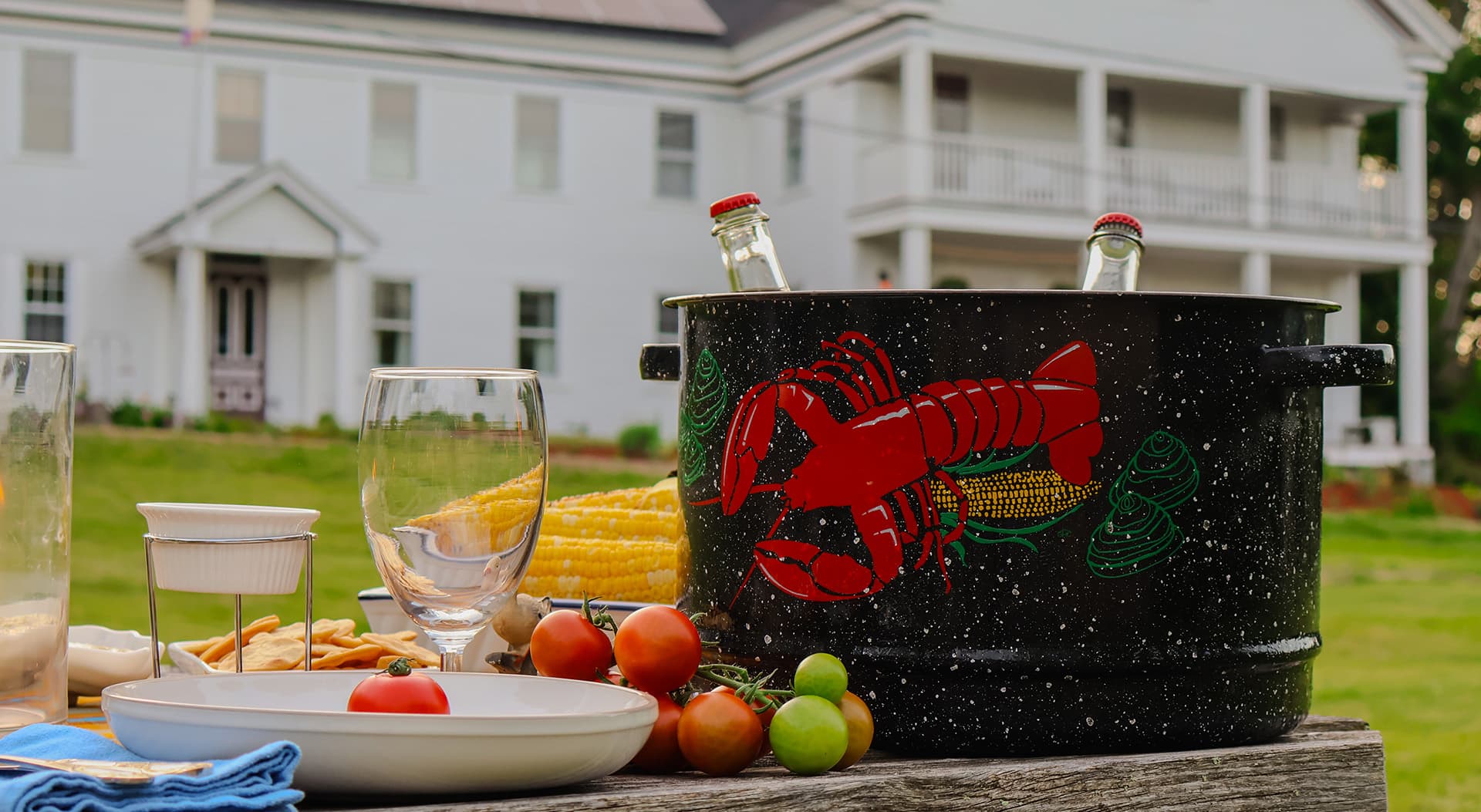 A large, decorative pot filled with lobsters sits on a wooden table next to glasses and plates, with a white house in the background.