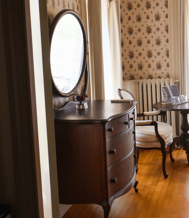 A wooden dresser with a round mirror, a small vase of flowers, and a table with a chair in a softly lit room with floral wallpaper.