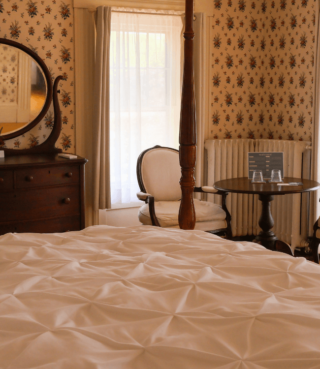 A cozy bedroom featuring a four-poster bed, antique furniture, and floral wallpaper.