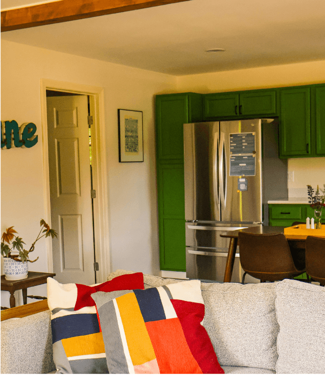 A cozy living space featuring a gray sofa with colorful pillows, a green kitchen, and a stainless-steel refrigerator.