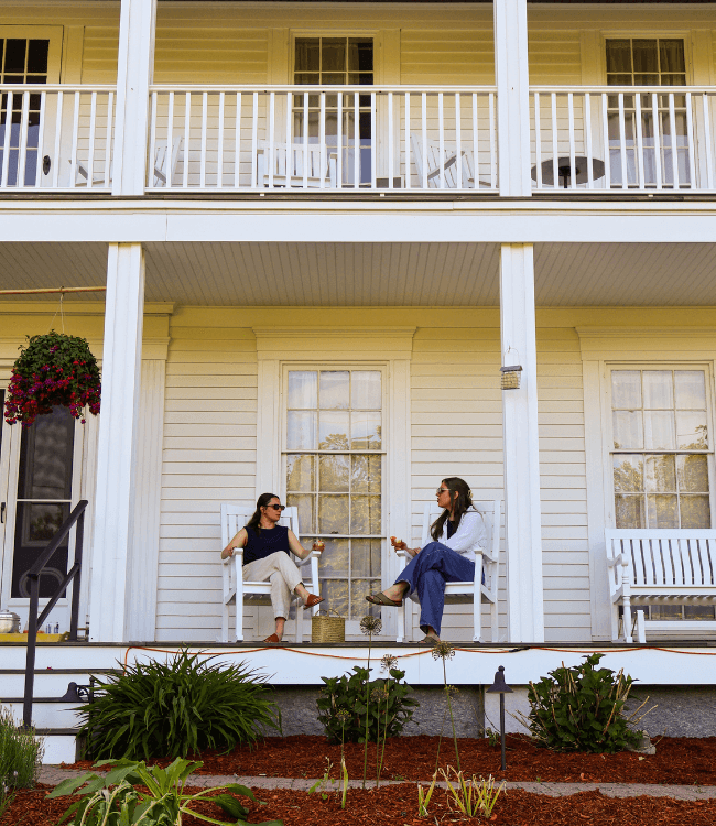 Two women sit smiling on a porch, enjoying drinks in a relaxed outdoor setting.